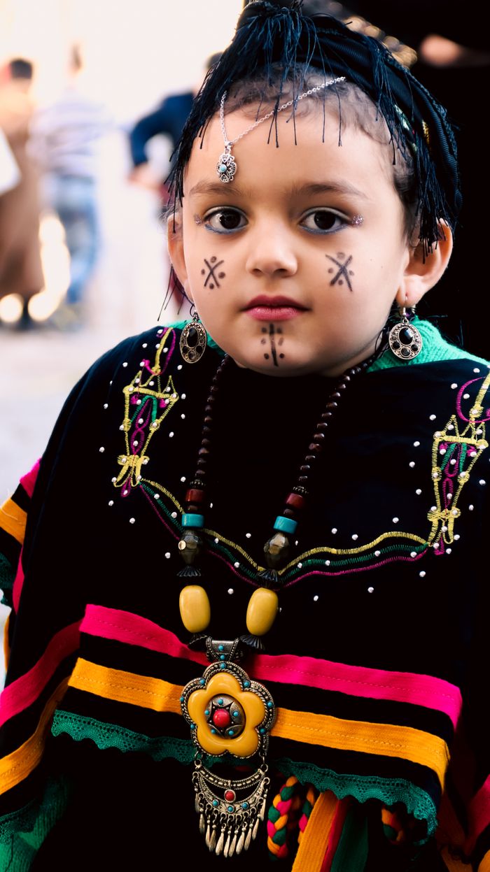 Captivating portrait of a child in colorful traditional Algerian clothing with face paint and intricate jewelry.