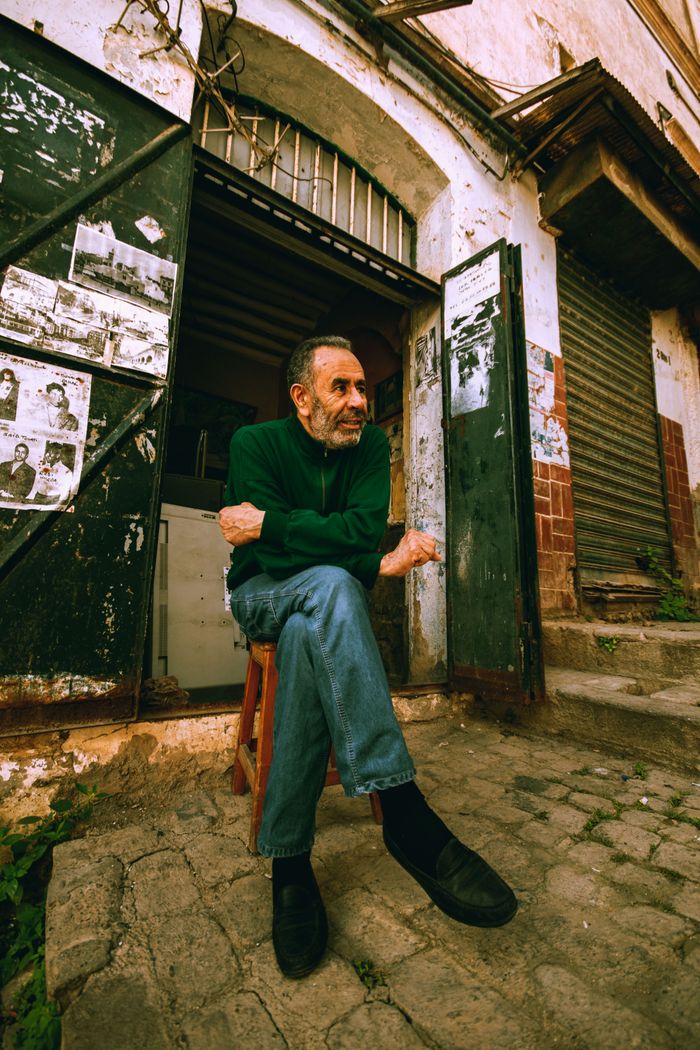 An elderly man in Algeria sitting on a chair outside a rustic shop, enjoying a day outdoors.
